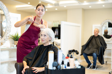 Senior woman is sitting in chair while cutting hair. Young woman master completes creation of...