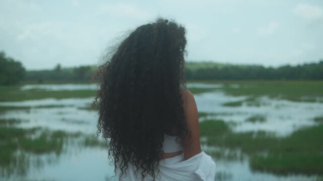 A woman with long, thick Type 3C curls and dark brown skin faces a flooded marshland under soft natural light, her hair gently lifted by the wind, evoking calm and introspection.