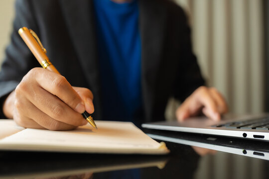 man sitting at a table at home working on a laptop and writing down ideas in a notebook.