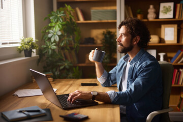 A man sits at a wooden home office desk, sipping coffee while focused on his laptop. The workspace...