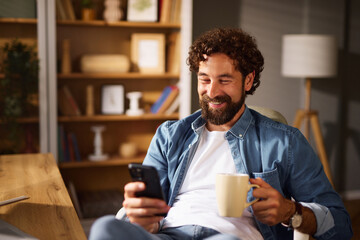 Smiling man relaxed in a comfortable chair, sipping coffee and browsing his phone in a well-decorated home office with bookshelves and warm lighting.
