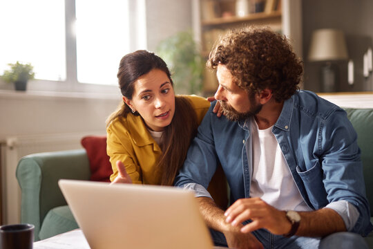 A couple is engaged in a serious conversation about their financial challenges while sitting together on a couch, focused on a laptop. The atmosphere suggests concern.