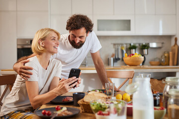 A couple shares a joyful moment over breakfast in their stylish kitchen, laughing together as they...