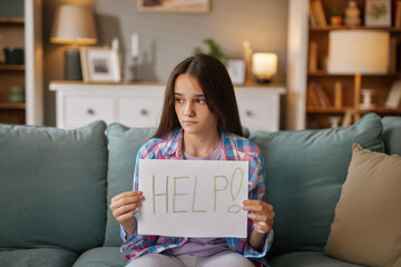 Young girl sits on a couch in a warm, inviting living room, holding a sign that reads help. She...