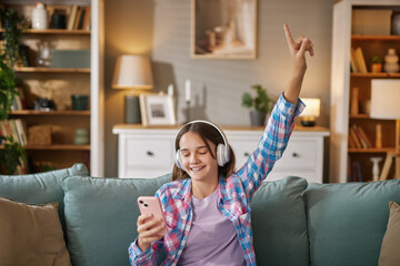 A teenage girl wearing headphones is joyfully listening to music in a living room, checking her...