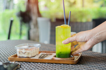 A person's hand holds a refreshing green tea sprinkled with fresh lemon with ice, some cake