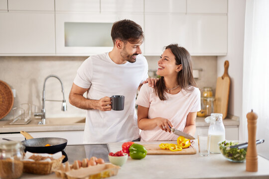Charming couple happily cooking together in a contemporary kitchen. The woman is chopping vegetables while the man holds a coffee mug, both sharing joyful moments and laughter. - Powered by Adobe