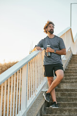 A man stands on outdoor stairs at sunset, dressed in sportswear and holding a water bottle,...