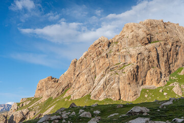 A majestic mountain slope with vast rock formations and patches of green grass at its base, under a bright blue sky with clouds