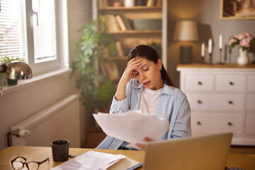 A woman sitting at a desk looks overwhelmed as she reviews financial documents in her tranquil home...