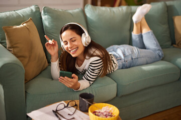 A woman lounges on a couch, wearing headphones and smiling as she listens to music on her phone....