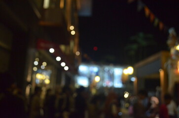 A dreamy blurred photo of a bustling night market, featuring warm lights, festive flags, and a crowd of indistinct shoppers, capturing a lively urban evening atmosphere.
