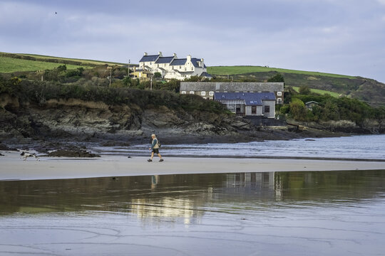 View of a lone figure strolling across a wet, sandy beach reflecting the sky, with coastal cottages nestled atop the rugged cliffs, Padstow, Cornwall, United Kingdom.