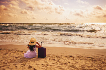Seascape during sunrise. A woman sits on the beach in summer with a travel suitcase. The girl looks...