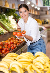 Smiling young woman purchaser holding tomato at the counter in big grocery store