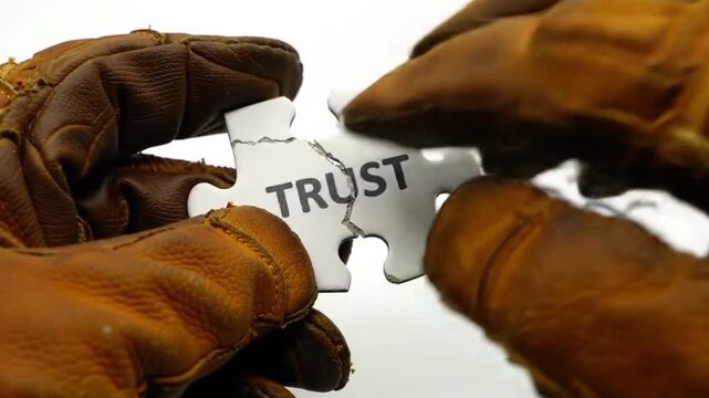 Two gloved hands carefully holding the two broken pieces of a white jigsaw puzzle spelling "TRUST" against a white background.