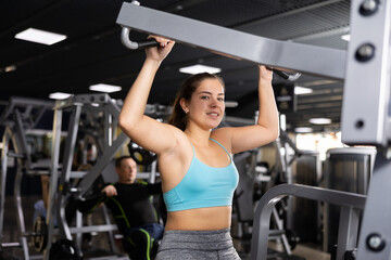 Fototapeta premium Young woman trains her arm muscles using a simulator in the gym