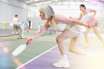 Focused active aged woman playing friendly pickleball match on indoor court. Senior people sports...