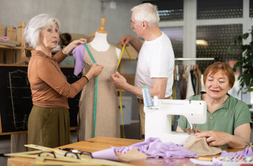 Elderly men and women spending time together sewing in cozy workroom, friendly chatting while using...