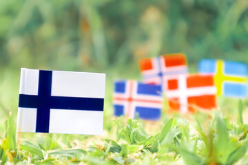 Finland national flag placed on green grass under sunlight, with blurred Nordic country flags in...