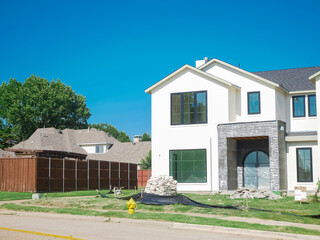 New construction home in well-established residential neighborhood suburbs Dallas, Texas, white house with bulk materials, erosion barrier, black framed windows, stone facade around entryway