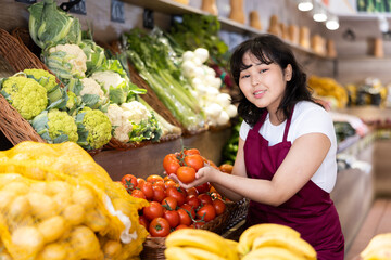 Cheerful friendly young asian salesgirl standing amount produce stands in greengrocery, holding...