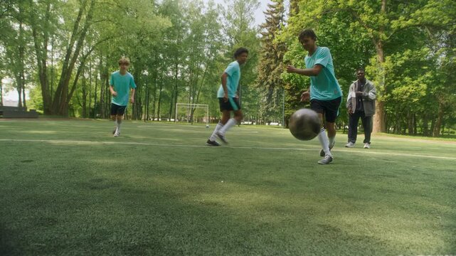 Three young diverse soccer players actively competing on grassy field while one of boys scoring goal toward camera, with Black male coach nearby observing and analyzing action during outdoor training