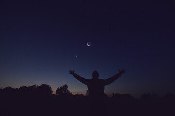 Silhouette of a man and countryside under the Moon, stars and planets in blue hour time.