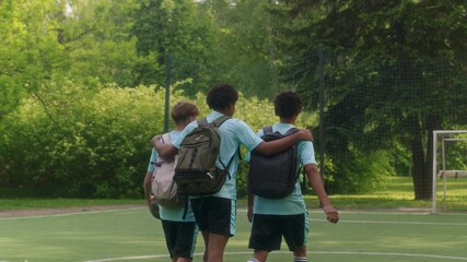 Back view shot of cheerful teen soccer player embracing two teammates from behind as they walking together toward outdoor field, wearing uniforms and backpacks on sunny day in park - Powered by Adobe