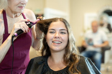 In beauty salon, cropped unrecognizable elderly woman hairdresser makes styling curls for young...
