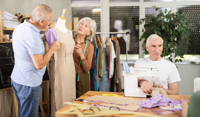 Senior couple talking while working with mannequin at sewing school studio, other male student...