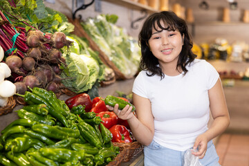 Positive interested young asian girl standing near shelves with ripe organic vegetables arranged in...