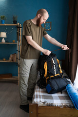 Young bearded man packing backpack on bed preparing for hiking trip in mountains in home interior
