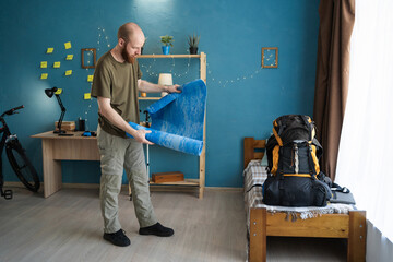 Young bearded hiker folding sleeping mat packing backpack for hiking in mountains at home