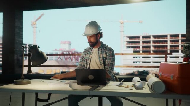 Construction engineer working on laptop at desk with refinery in background