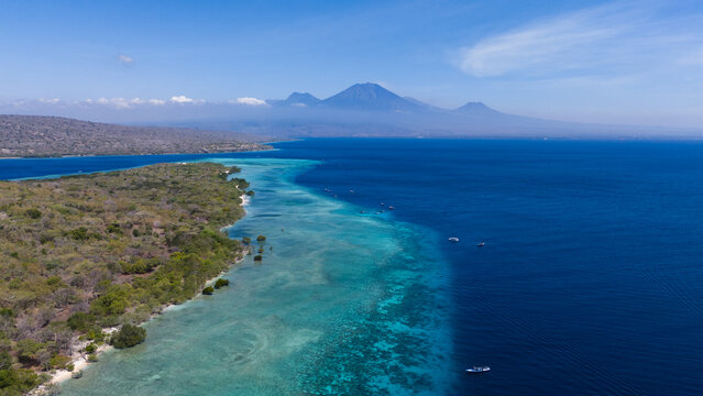 Aerial view of where the turquoise shallows meet the deep blue sea near the lush green shores, Mt. Ijen volcano on the island of Java in the background, Menjangan Island, Bali, Indonesia.