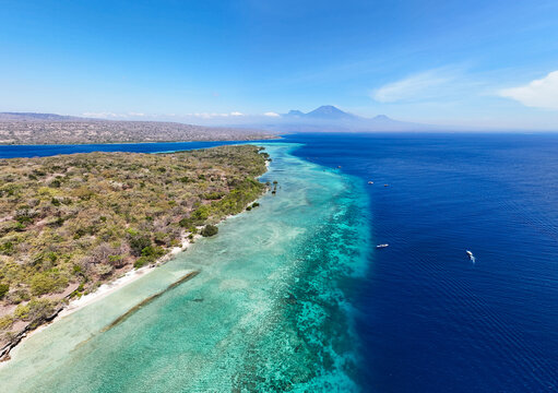 Aerial view of where the turquoise shallows meet the deep blue sea near the lush green shores, Mt. Ijen volcano on the island of Java in the background, Menjangan Island, Bali, Indonesia.