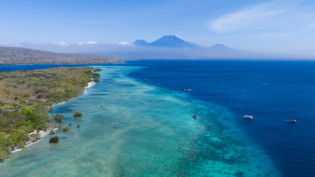 Aerial view of where the turquoise shallows meet the deep blue sea near the lush green shores, Mt. Ijen volcano on the island of Java in the background, Menjangan Island, Bali, Indonesia.