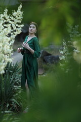 A woman in a flowing emerald green dress poses amidst white blossoms and lush garden foliage,...