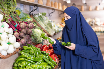 Portrait of girl in paranja buying fresh bell peppers in grocery shop