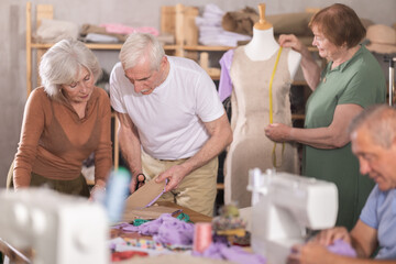 Small group of senior learners enjoying relaxed sewing class in cozy workshop, measuring,...