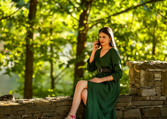 A woman in an emerald green dress sits on a stone wall under dappled golden-hour light, holding her...