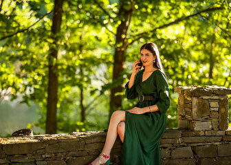 A woman in an emerald green dress sits on a rustic stone wall in golden light, holding her phone to...