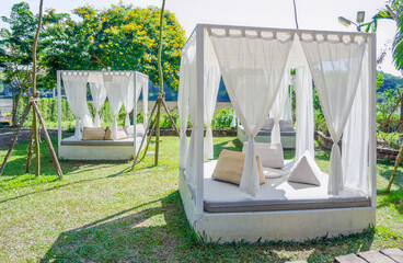 Luxurious White Daybeds with Drapes in a Serene Outdoor Garden Setting. White daybeds with sheer drapes and plush cushions are set on sunlit lawn, surrounded by trees, offering relaxing outdoor escape