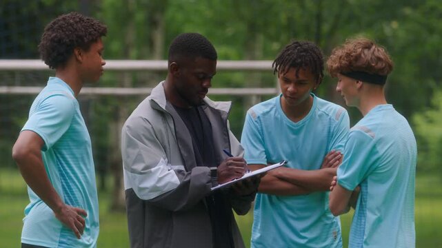 Medium shot of diverse group of young soccer players gathering around their coach on outdoor field as he drawing game plan on clipboard during game strategy and tactics discussion