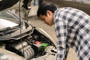 Asian man examining the engine of a white car with the hood open, appearing focused and concerned...