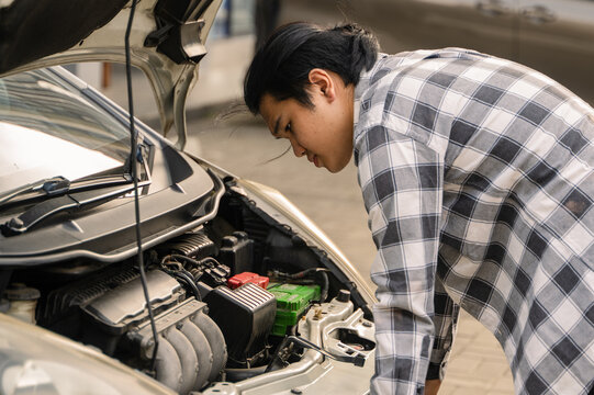 Asian man examining the engine of a white car with the hood open, appearing focused and concerned while inspecting under bright daylight in an outdoor setting - Powered by Adobe