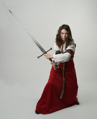 portrait of pretty young female brunette model wearing a red medieval historical gown holding fantasy sword weapon. standing pose, Isolated figure on white studio background.