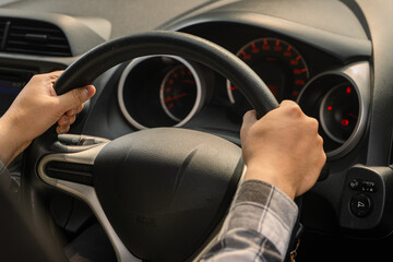 Close-up of an Asian person’s hands holding the steering wheel of a car from the driver’s seat,...