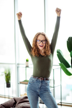Fototapeta Joyful young woman celebrating achievement indoors while raising arms with a happy expression surrounded by modern interior